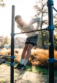 Sportiškai atrodantis vyras atlieka prisitraukimą ant juodo metalinio stovo, vilkėdamas baltą ilgomis rankovėmis marškinį, pilkas šortus ir juodus sportinius batus su baltais kojiniais.