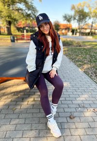 Colete acolchoado preto sobre um moletom cinza, leggings roxas e ténis brancos com detalhes em pastel. Usando um boné preto com o logo "LA".