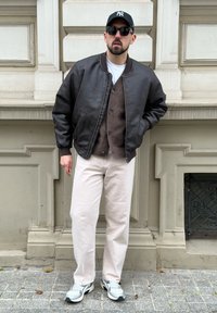 Mees mustas nahktagus, pruunis kardiganis, valgetes pükstes, valgetes tossudes, musta mütsi ja prillidega seismas beežist kiviseinast.