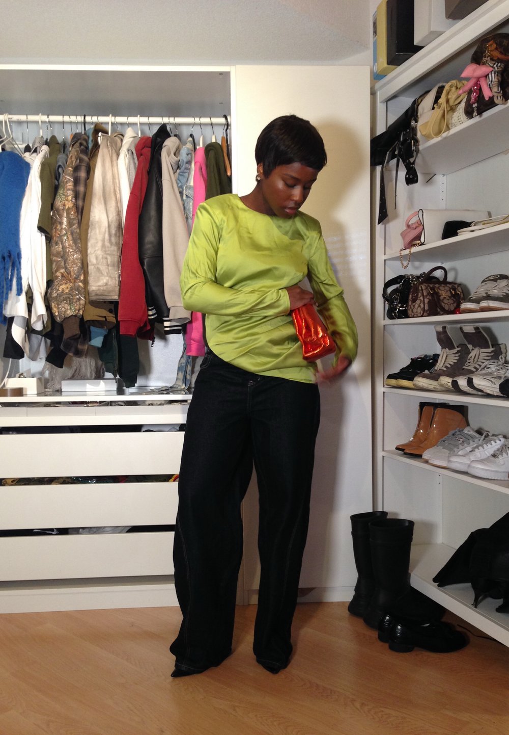 Camisa verde de mangas longas com uma textura suave, combinada com jeans azul escuro. O sujeito segura uma clutch laranja. Um guarda-roupa é visível ao fundo.