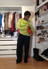 Camisa verde de mangas longas com uma textura suave, combinada com jeans azul escuro. O sujeito segura uma clutch laranja. Um guarda-roupa é visível ao fundo.