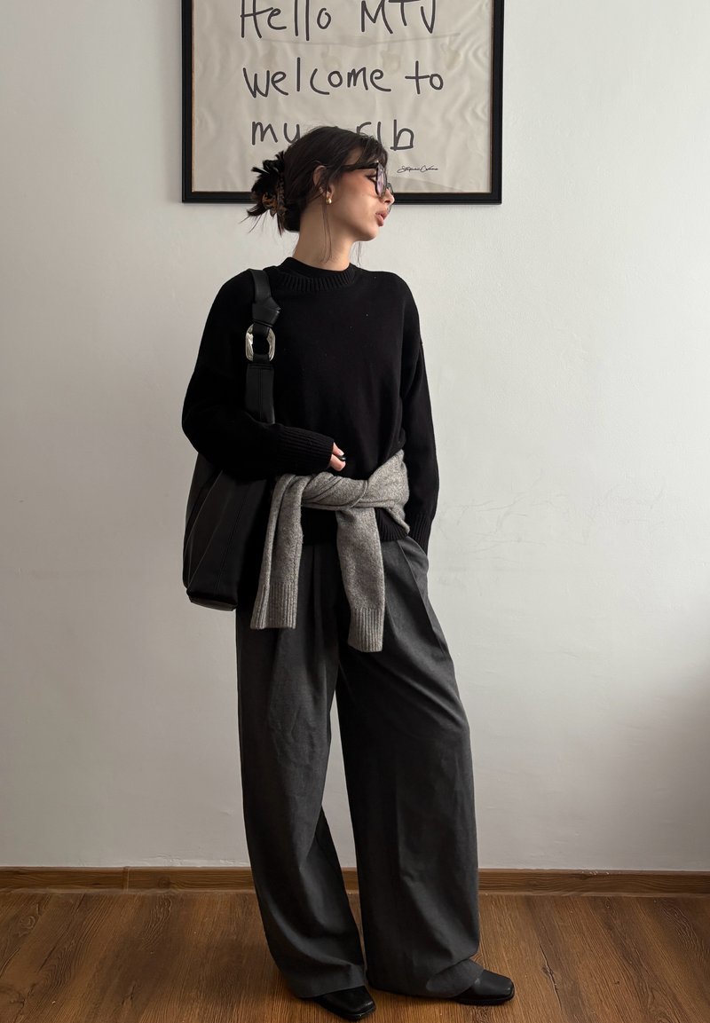 Femme en pull noir et pantalon large gris se tient à l'intérieur sous un panneau encadré avec l'inscription "Hello MTV bienvenue dans mon club."