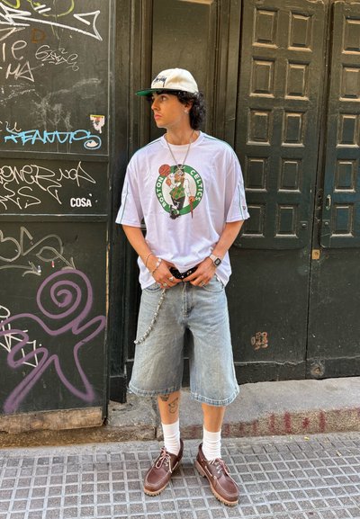 Joven con camiseta de los Boston Celtics, pantalones cortos de mezclilla, calcetines blancos, zapatos marrones y gorra blanca, de pie frente a una pared de graffiti y puertas de madera oscura.