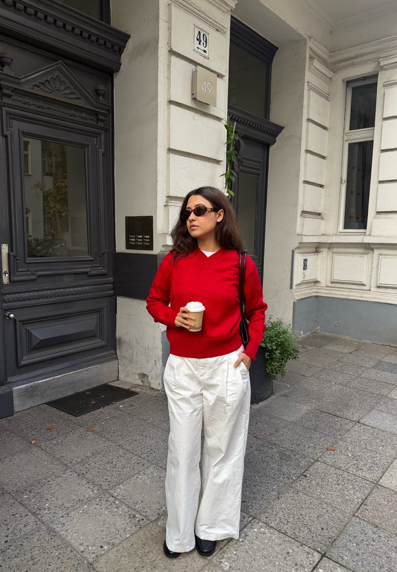 Femme portant un pull rouge et un pantalon blanc, tenant une tasse de café, debout sur un trottoir près d'une entrée de bâtiment numérotée 49.