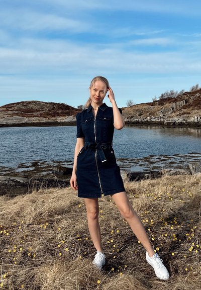 Vestido camisero de denim oscuro con cremallera en la parte delantera, mangas cortas, cintura ajustada y bolsillos, combinado con zapatillas blancas sobre hierba con flores amarillas.