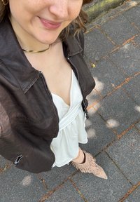 Jaqueta de couro castanha, vestido branco com tecido drapeado e botas de tornozelo bege com franjas. O fundo apresenta pavimento cinzento com folhas espalhadas.