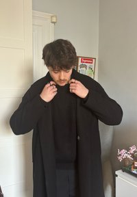 Ung mann med brunt hår som justerer kragen på en lang svart frakk innendørs, stående ved en hvit dør og et bord med en liten blomsterdekorasjon.