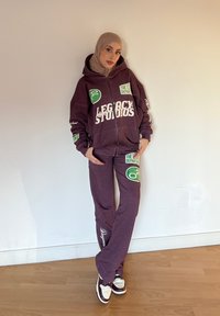 Sweatshirt com fecho, de cor bordeaux, com logótipos gráficos, emparelhado com calças de fato coordenadas. As calças apresentam detalhes impressos e um ajuste confortável. Ténis castanhos completam o visual.
