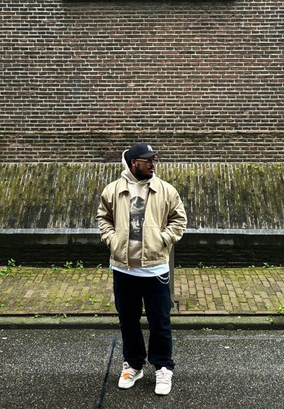 Chaqueta beige sobre una sudadera gris con un gráfico. Pantalones negros y zapatillas de colores claros. De pie contra una pared de ladrillo.