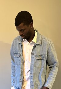 Veste en denim bleu clair avec deux poches avant, portée par-dessus une chemise blanche à boutons. Le modèle a les cheveux noirs courts et texturés.