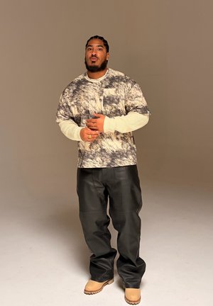 Hombre con trenzas llevando una camisa estampada de gran tamaño sobre una camiseta blanca de manga larga, pantalones de cuero negro y botas color canela, de pie frente a un fondo liso.