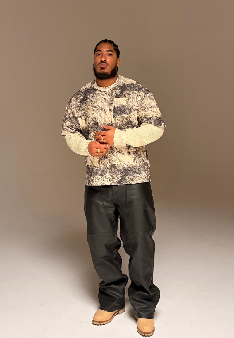 Homme aux cheveux tressés portant une chemise imprimée oversize par-dessus un t-shirt blanc à manches longues, un pantalon en cuir noir et des bottes couleur fauve, debout devant un fond uni.