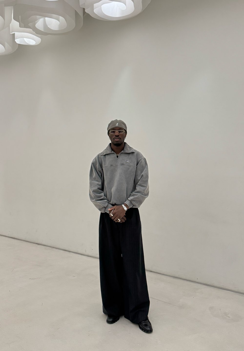 Grauer Fleece-Pullover mit Reißverschluss, weite schwarze Hose und dunkle Schuhe. Accessoires sind runde Brille und passende Mütze. Minimalistischer Hintergrund.