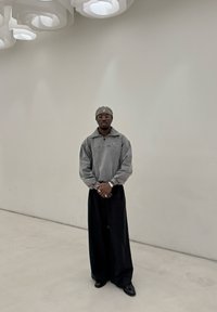 Grauer Fleece-Pullover mit Reißverschluss, weite schwarze Hose und dunkle Schuhe. Accessoires sind runde Brille und passende Mütze. Minimalistischer Hintergrund.