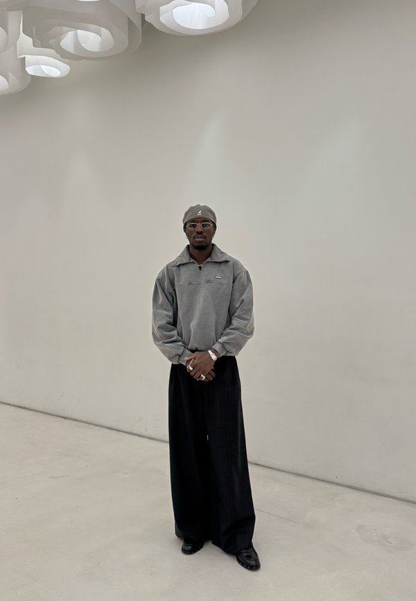 Grauer Fleece-Pullover mit Reißverschluss, weite schwarze Hose und dunkle Schuhe. Accessoires sind runde Brille und passende Mütze. Minimalistischer Hintergrund.