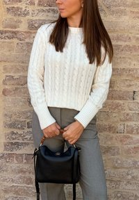 Pulover crem tricotat cu model cable, cu manșete cu nervuri, pantaloni gri tailored și o geantă de mână din piele neagră cu mâner din bambus și detalii din aur.