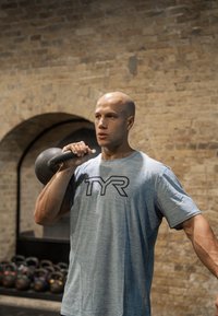 Kettlebell, der von einer Person mit einem hellblauen Sportshirt mit "TYR"-Logo in einer Hand gehalten wird, vor einem Ziegelmauer-Hintergrund.