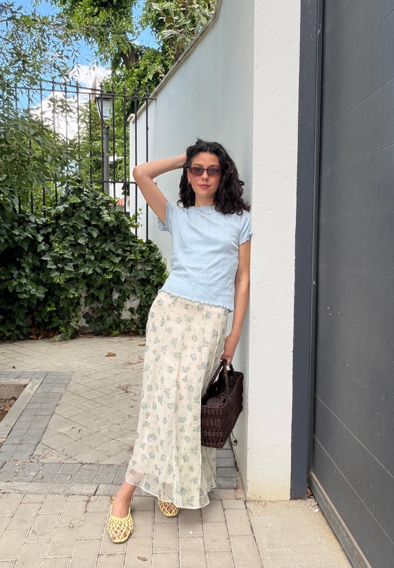 Femme aux cheveux bouclés portant des lunettes de soleil, un haut bleu à manches courtes, une jupe fleurie, des chaussures jaunes, tenant un sac tressé, appuyée contre un mur blanc à l'extérieur.
