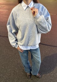 Sweat-shirt gris à manches raglan porté sur une chemise blanche à col, associé à un jean large bleu et des bottines marron foncé. Tissu texturé.