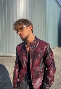 Homme aux cheveux bouclés portant des lunettes de soleil teintées, un collier en chaîne argentée et une veste en cuir bordeaux, se tenant à l'extérieur près d'un mur en métal ondulé.