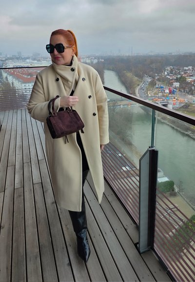 Mujer de cabello rojo vestida con un abrigo beige y botas negras, de pie en un balcón con vistas a un río y un paisaje urbano en un día nublado.