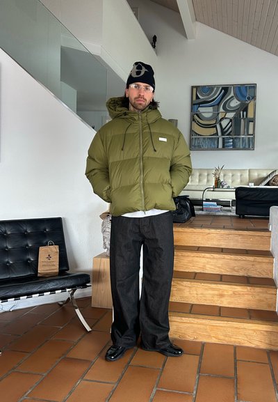 Hombre con chaqueta acolchada verde oliva, gorro negro, gafas y pantalones anchos oscuros de pie en el interior cerca de una escalera de madera y muebles modernos.