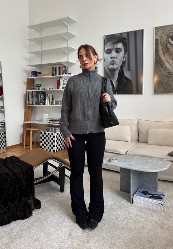 Žena v šedém svetru na zip a černých kalhotách držící černou tašku, stojící v moderním obývacím pokoji s monochromatickým uměním a nábytkem.