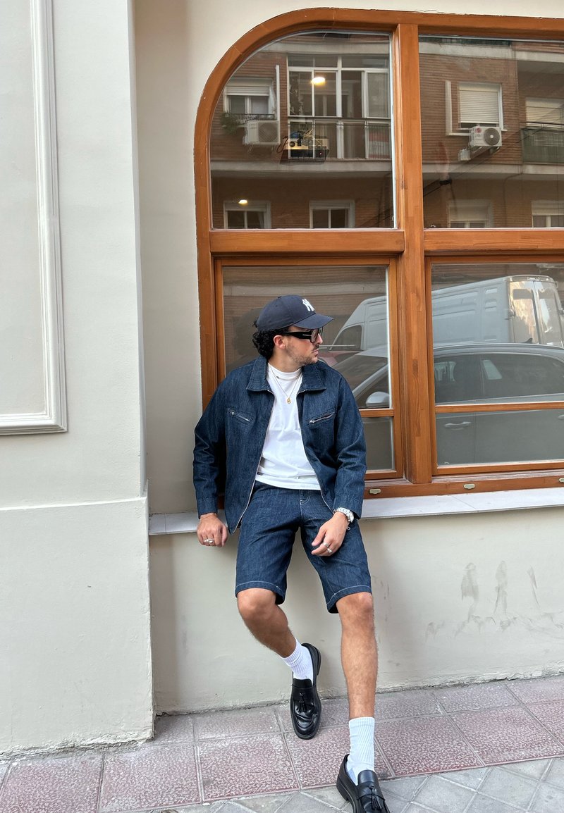 Casaco de denim e calções de ganga de um tom escuro, t-shirt branca por baixo, sapatos pretos, meias brancas, sentado junto a uma janela com moldura de madeira.
