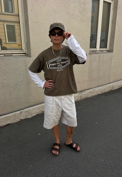 Persona con gafas de sol, una gorra, camiseta marrón sobre una camisa blanca de manga larga, pantalones cortos blancos y sandalias, de pie en una calle cerca de una pared de un edificio.