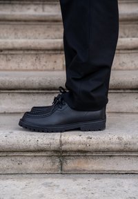 Scarpe in pelle nera con design mocassino, cuciture testurizzate e suole in gomma spesse, esposte su gradini di pietra.