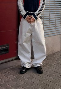 Personne portant un pantalon blanc trop grand, des chaussures noires et un pull bleu marine avec des manches blanches et rouges, debout sur un trottoir pavé.