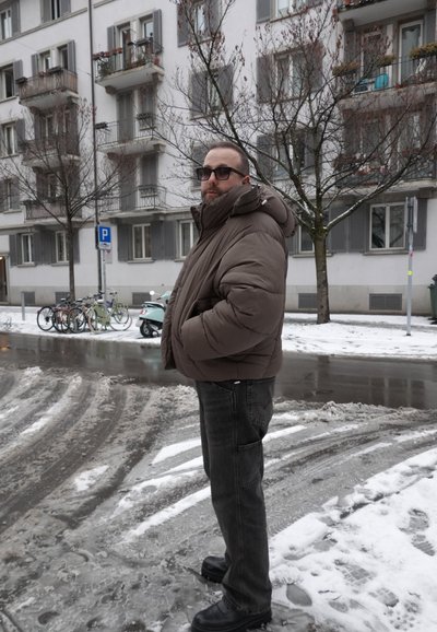 Hombre con chaqueta acolchada marrón, gafas de sol y pantalones negros de pie en una calle nevada cerca de bicicletas aparcadas y edificios residenciales.