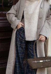 Manteau en laine beige sur un pull beige clair, assorti à un pantalon à carreaux bleu marine. Tenant un sac à main marron foncé avec une bandoulière à accents dorés.