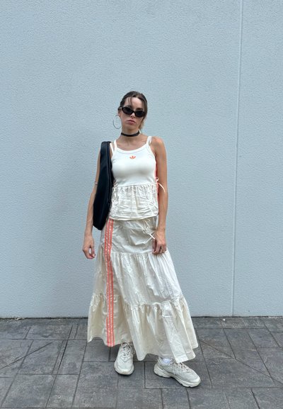 Top blanco sin mangas con logo rojo, falda crema con capas y rayas rojas, bolso negro de gran tamaño y zapatillas blancas gruesas; gafas de sol y choker.