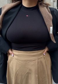 Sort ribbet top med et lille rødt logo, parret med en beige nederdel med plisseringer og en sort blazer med et brunt uldhalstørklæde drapere over.