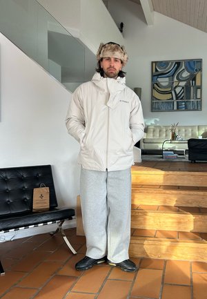 Hombre con chaqueta acolchada blanca, pantalones deportivos grises, zapatos negros y gorro de piel de pie en el interior junto a unas escaleras y muebles modernos.