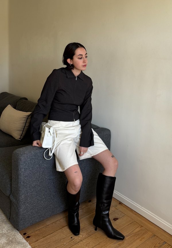 Jacket cropped preta com fecho e manga longa, calções brancos e botas altas de pele pretas. Pequena mala branca repousa no sofá.