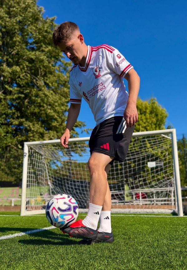 Hombre con camiseta blanca del Liverpool controla el balón de fútbol cerca del poste de la portería en un campo verde bajo un cielo azul claro.