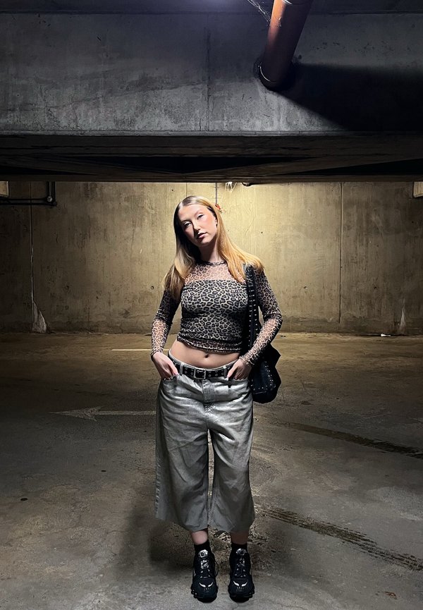 Młoda kobieta o długich blond włosach stoi z rękami w kieszeniach w słabo oświetlonym betonowym garażu, ubrana w bluzkę w panterkę i szerokie spodnie.