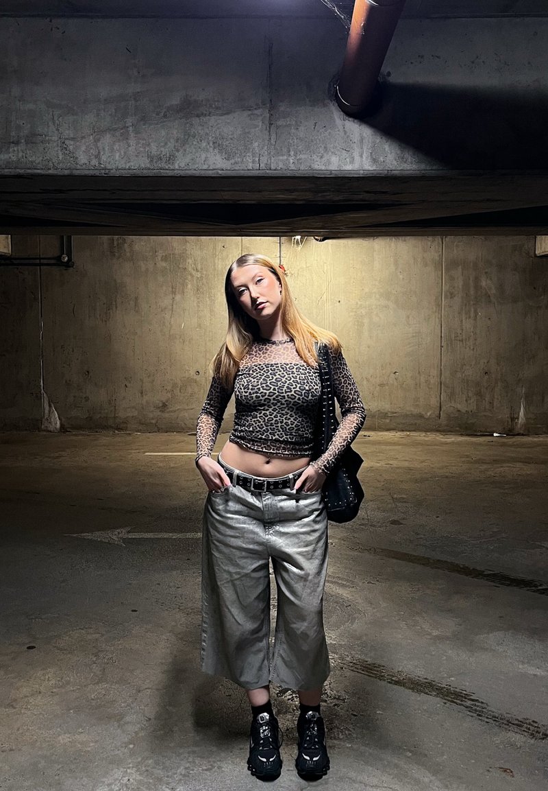 Jeune femme aux longs cheveux blonds, les mains dans les poches, dans un parking en béton faiblement éclairé, portant un haut à motifs léopard et un pantalon à jambes larges.