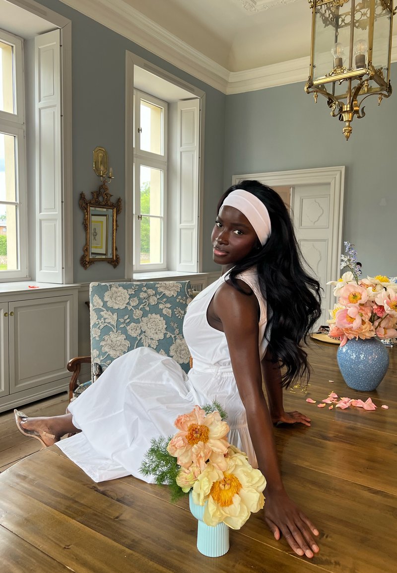 Femme en robe blanche et bandeau assise à une table en bois avec des vases à fleurs dans une pièce lumineuse et ornée avec de grandes fenêtres et des murs bleus.