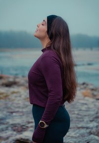 Jeune femme aux longs cheveux bruns, portant un haut violet et un bandeau noir, debout en plein air près de l'eau, les yeux fermés et la tête inclinée en arrière.