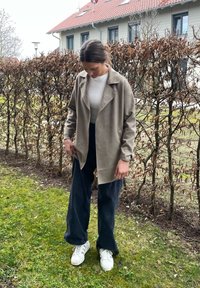 Beige, könnyű dzseki, hajtókával és nyitott elülső résszel, sötét, bőszárú nadrággal és fehér tornacipővel a füvön, kültéren, sövények mellett.