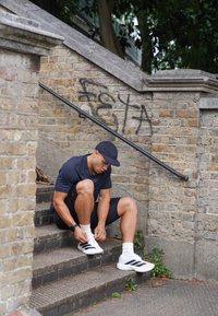 Mężczyzna w czarnym ubraniu sportowym siedzi na schodach na świeżym powietrzu, wiążąc swoje białe trampki w czarne paski, obok ceglanej ściany z graffiti.