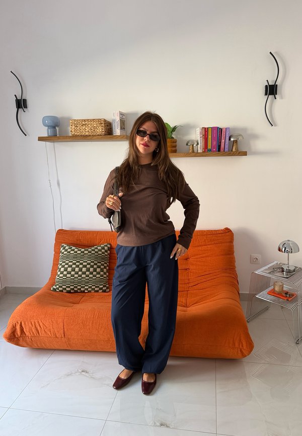 Mujer con gafas de sol, camiseta marrón, pantalones azules y zapatos marrones de pie frente a un sofá naranja con un cojín a cuadros en una sala de estar moderna.