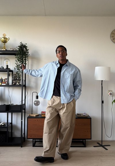 Joven con gafas, camisa azul claro a cuadros, camiseta negra y pantalones beige de pie en la sala de estar con estanterías, lámpara y armario de madera.