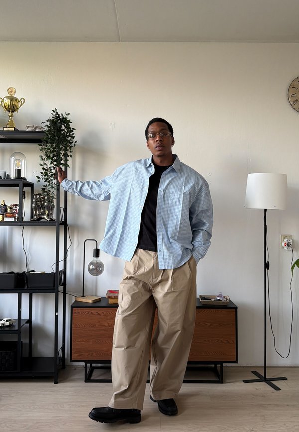 Jeune homme portant des lunettes, une chemise à carreaux bleu clair, un t-shirt noir et un pantalon beige debout dans un salon avec des étagères, une lampe et un meuble en bois.