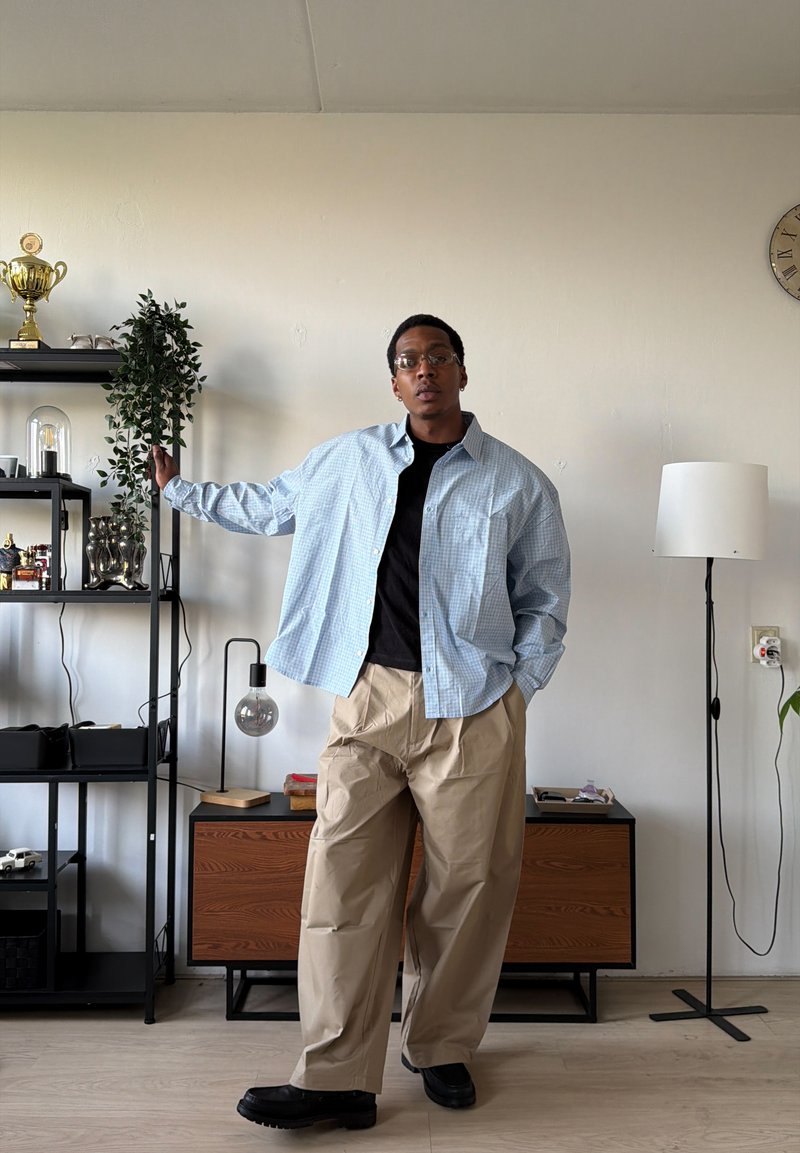 Jeune homme portant des lunettes, une chemise à carreaux bleu clair, un t-shirt noir et un pantalon beige debout dans un salon avec des étagères, une lampe et un meuble en bois.