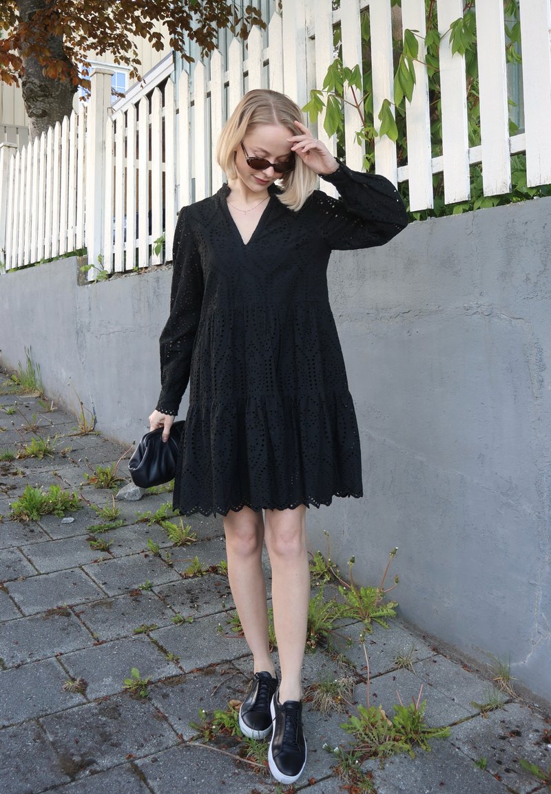 Femme en robe noire et lunettes de soleil se tient sur un trottoir près d’une barrière blanche, tenant un sac noir et touchant ses cheveux.