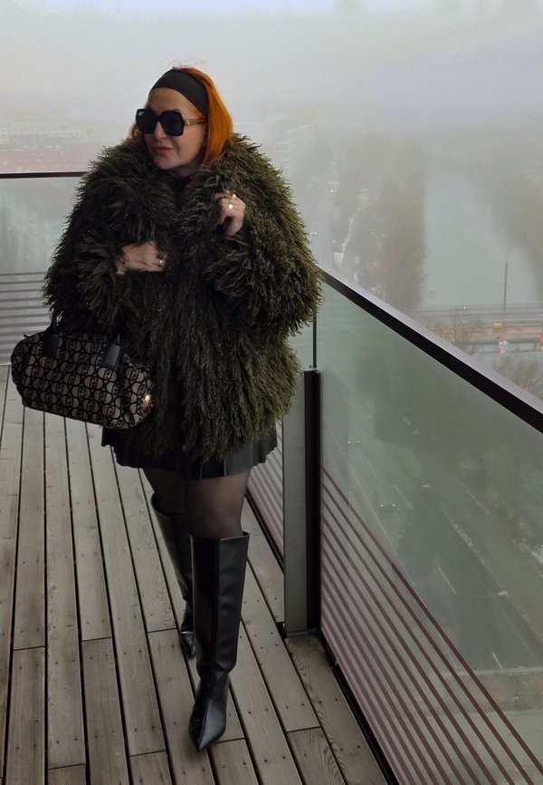 Mujer con cabello naranja, gafas de sol oscuras, abrigo grande de piel verde, falda negra y botas, sosteniendo un bolso con estampado en un balcón con niebla.